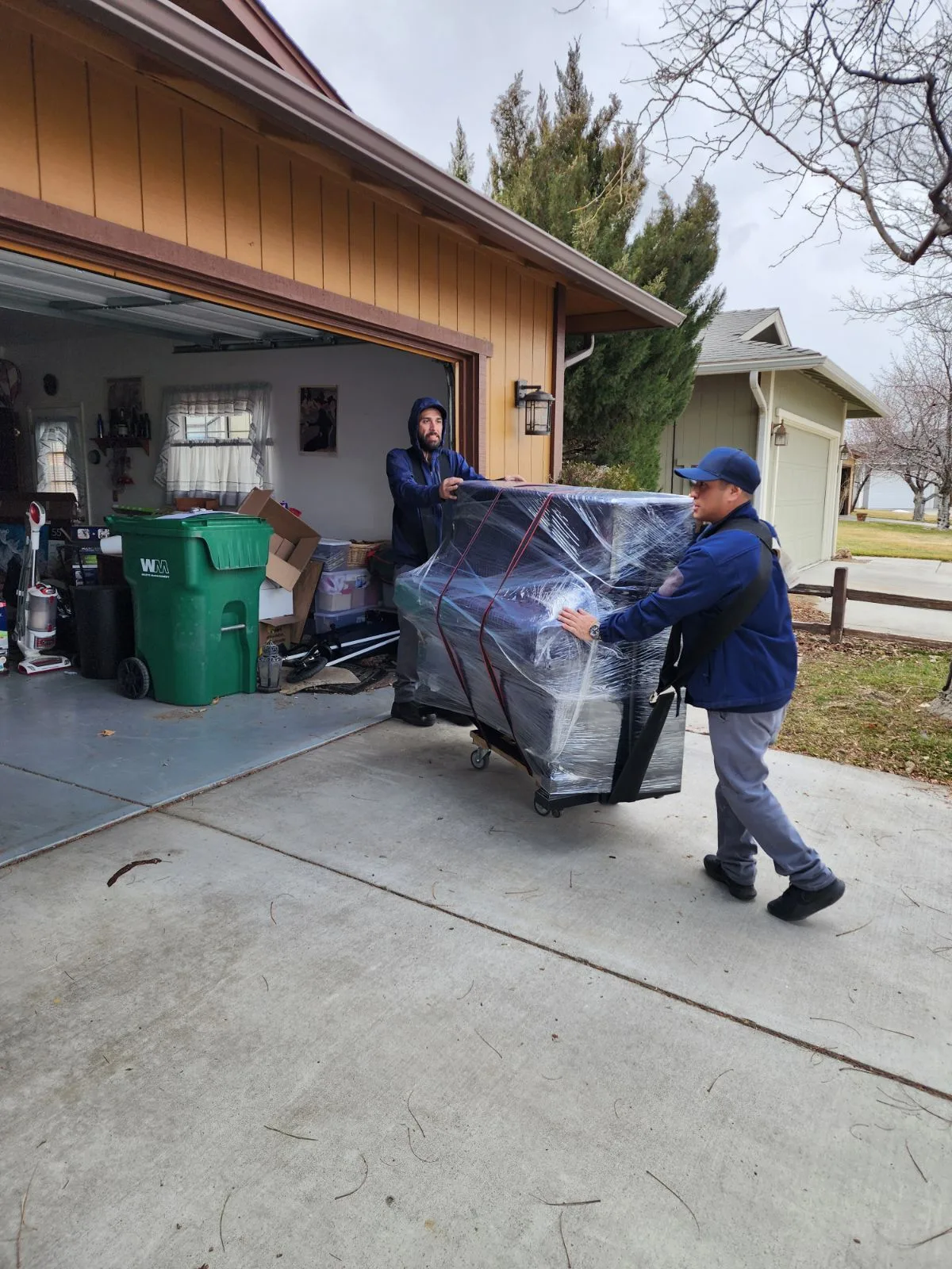 Two Green Echo movers loading a shrink-wrapped piece of furniture from a garage onto a dolly